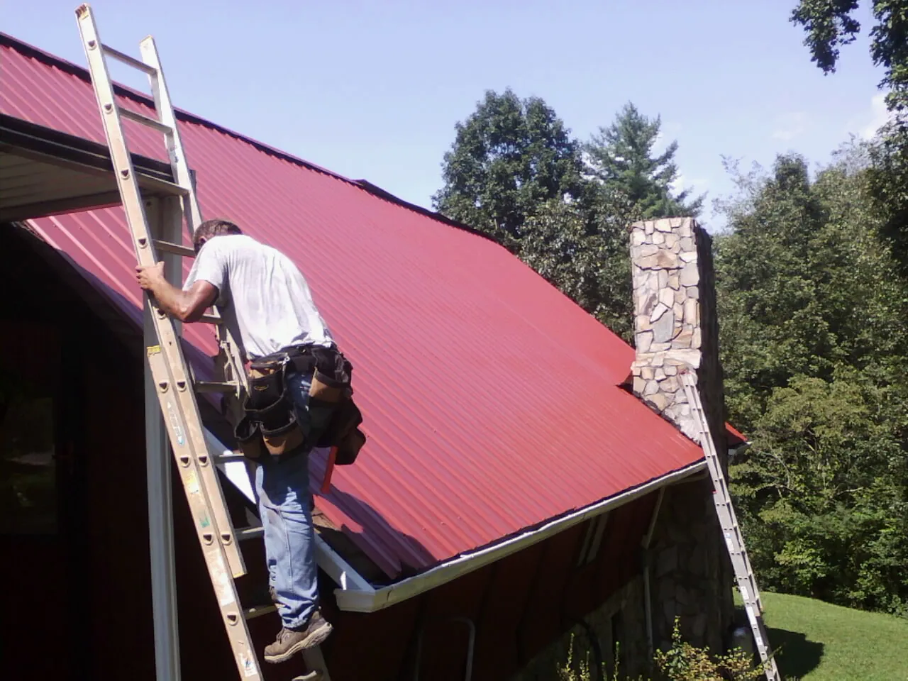 Roofer on a ladder beside a red metal roof and stone chimney — Soffit & Fascia Repair by Roofing Brothers in Newmanstown